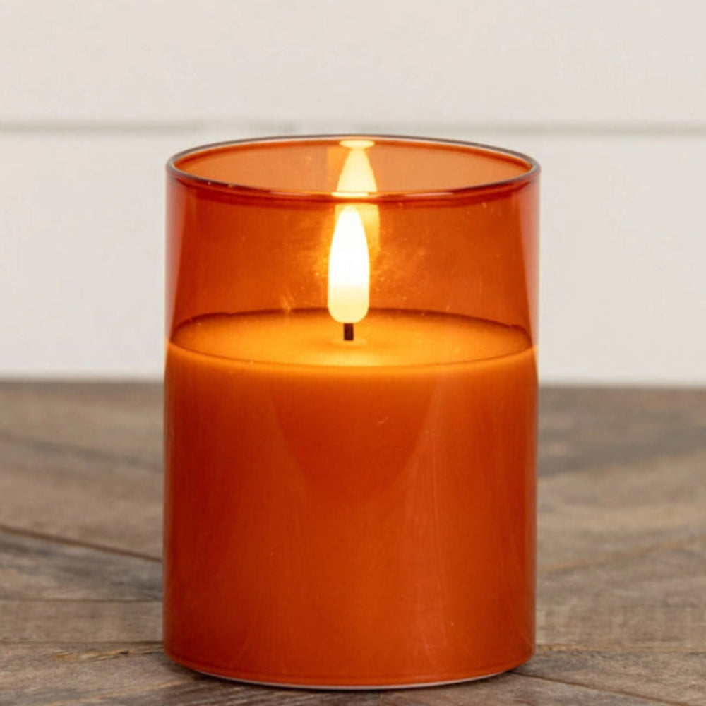 Lit flameless orange candle inside a sienna glass cylinder resting on a wooden surface.