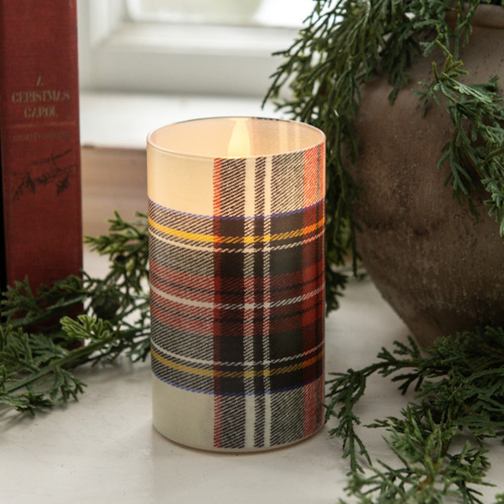 Lit candle in a white tartan glass holder next to evergreen greenery and a red book.