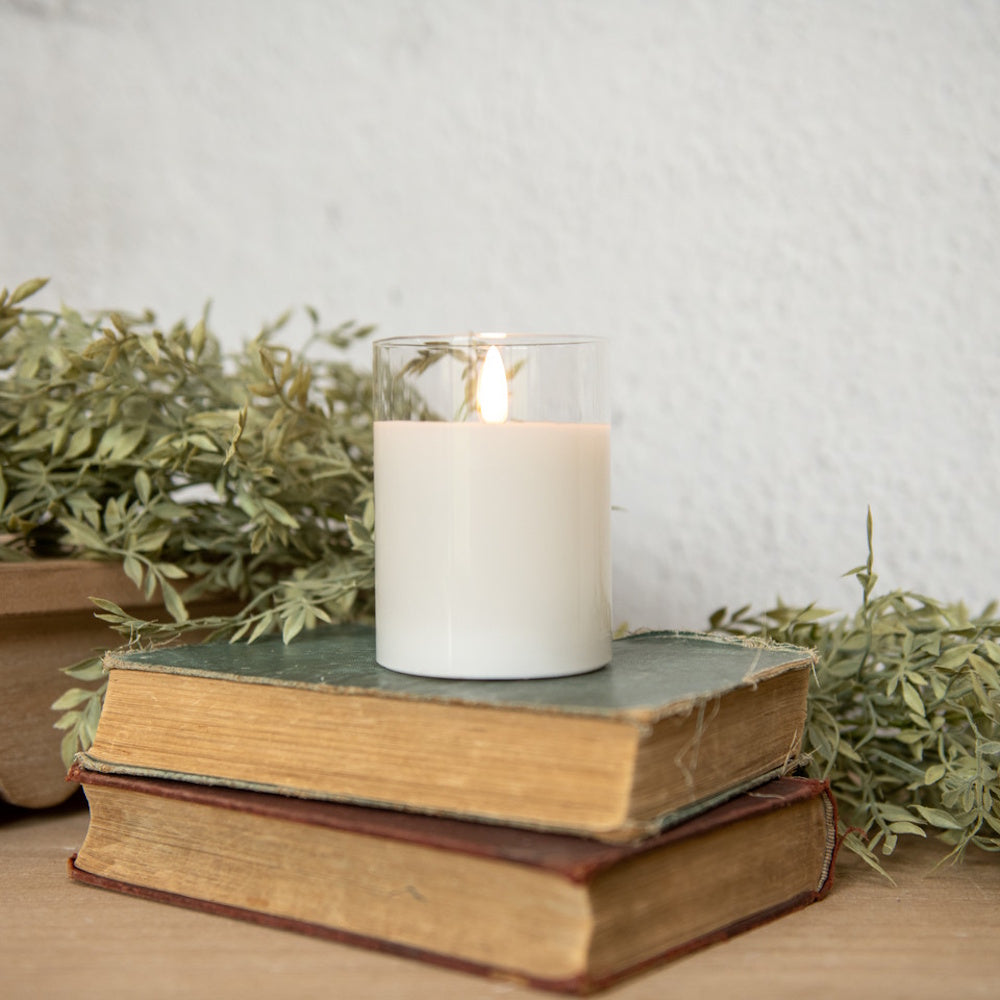 A glowing white candle in a clear glass holder sitting on stacked vintage books with leafy greenery nearby.
