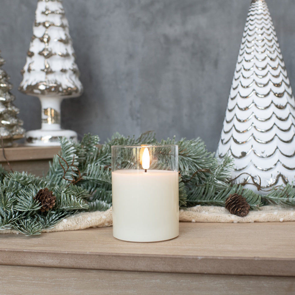 White glass flameless candle set among pine greenery, pinecones, and decorative white Christmas trees.