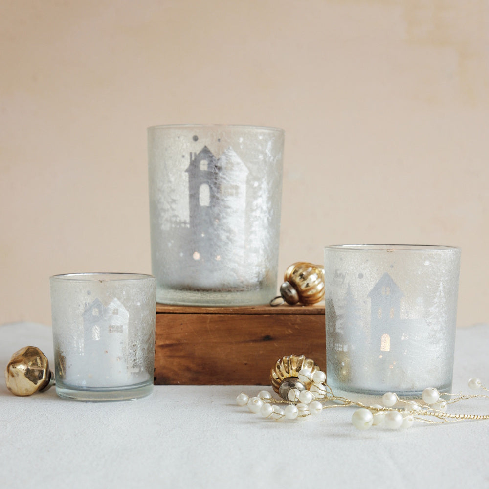 Silver frosted glass votive holders etched with winter house scenes, surrounded by festive ornaments.