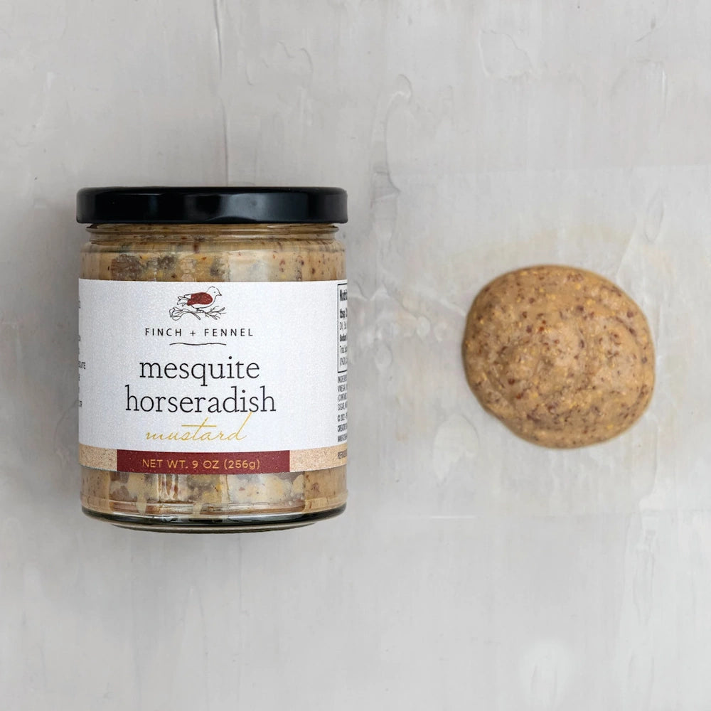 Finch + Fennel mesquite horseradish mustard jar and a dollop of grainy mustard on a light grey background.