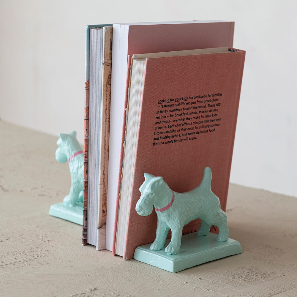 Mint-green metal dog bookends holding up three books, including a pink cookbook, on a textured surface.