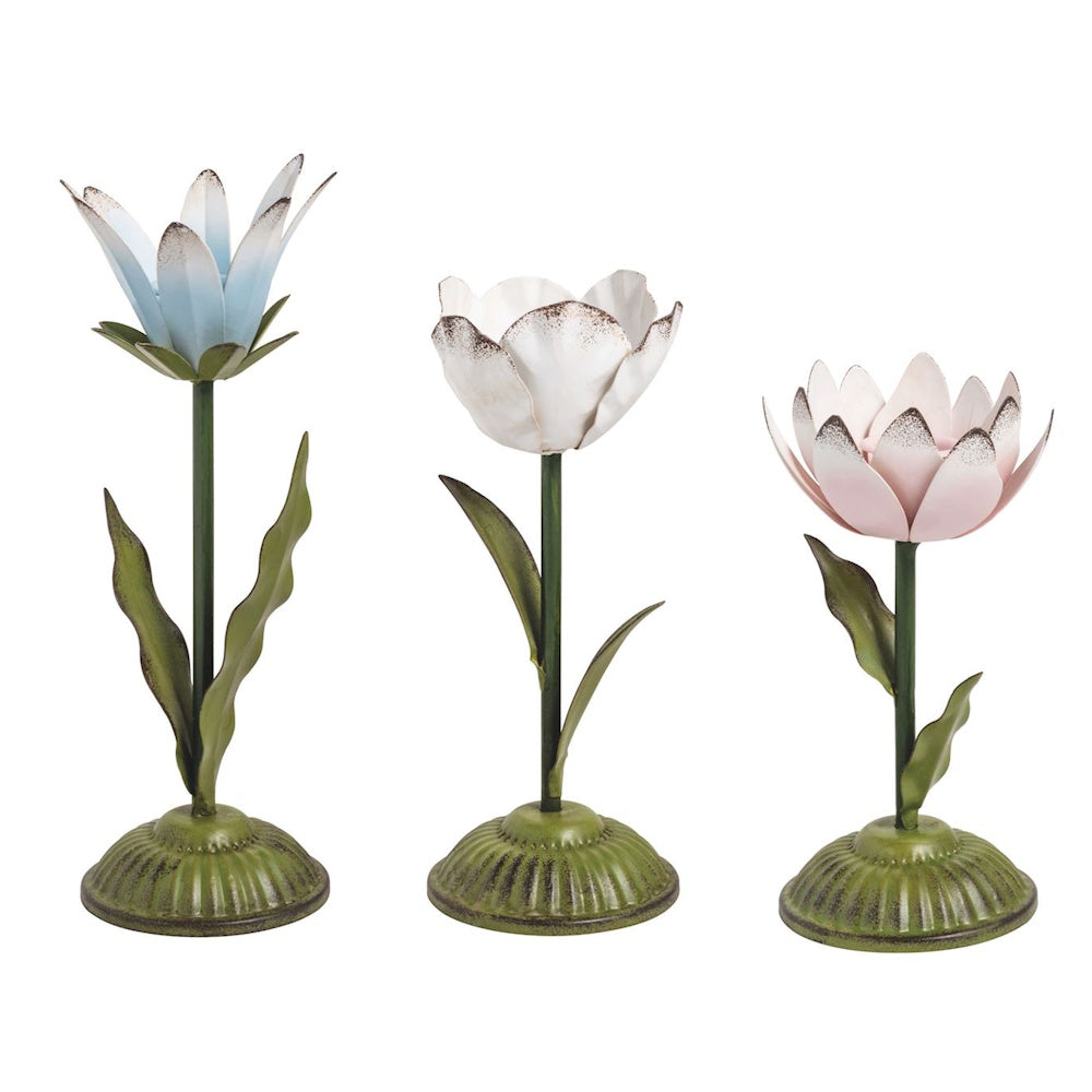 Three metal flower-shaped candlesticks in blue, white, and pink with green stems and bases on a white background.