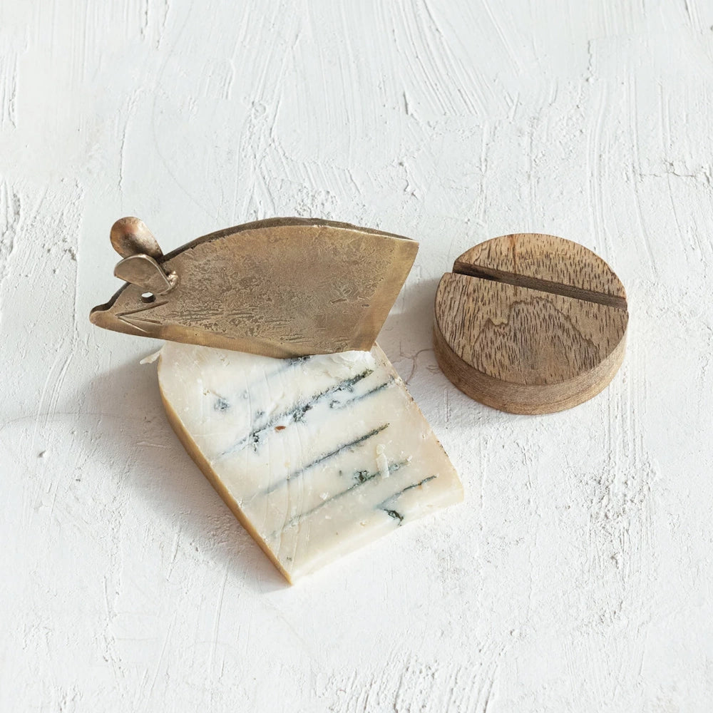 Brass mouse-shaped cheese knife slicing blue cheese next to a round mango wood base on a white textured surface.