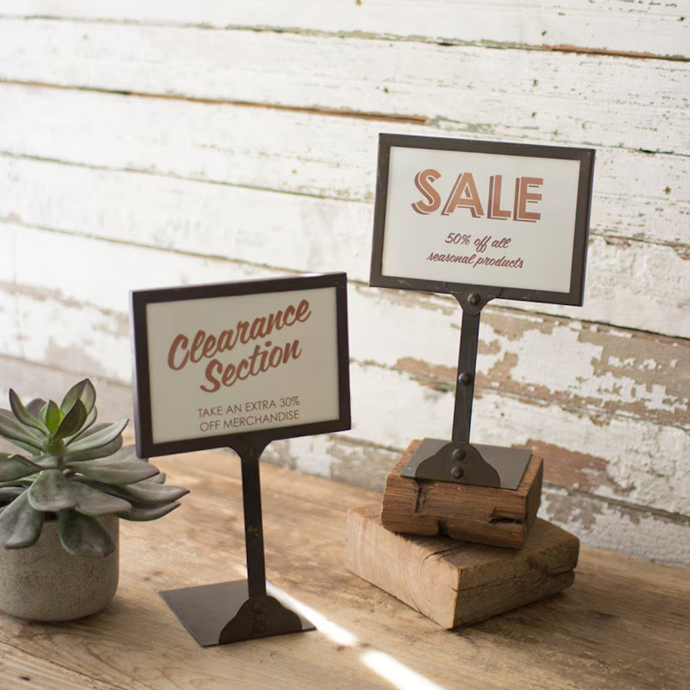 Two metal tabletop sign holders with dark frames, displaying sale and clearance promotional signs.