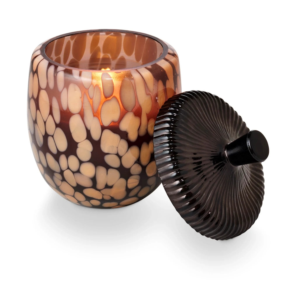 Lit candle in a brown tortoiseshell speckled glass jar with a dark, ribbed lid leaning beside it.