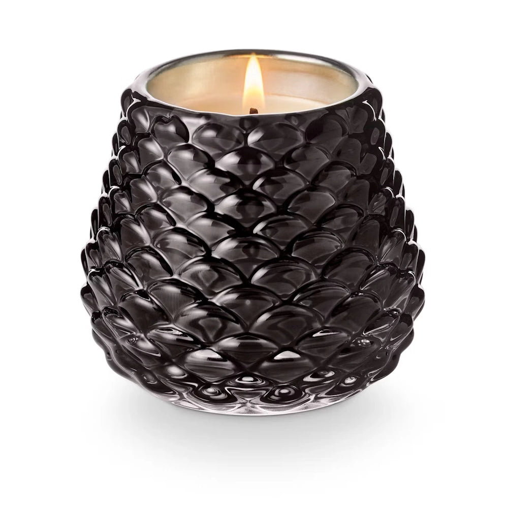 Lit votive candle inside a glossy black, textured pinecone-shaped glass holder on a white background.