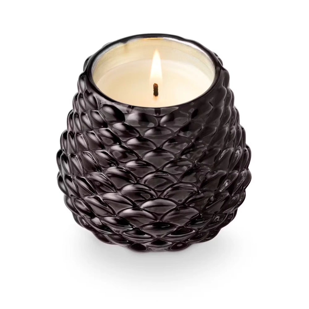 Lit scented candle in a textured glossy black glass pinecone votive holder.