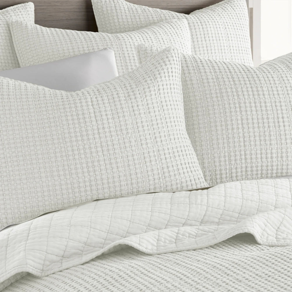 White waffle-weave quilt and matching pillows layered on a bed with a textured, modern aesthetic.