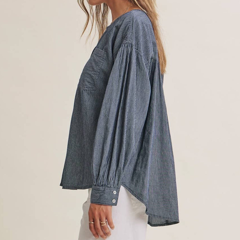 Side profile of a long-sleeved, pinstriped blue chambray henley top with gathered sleeves and a high-low hem.