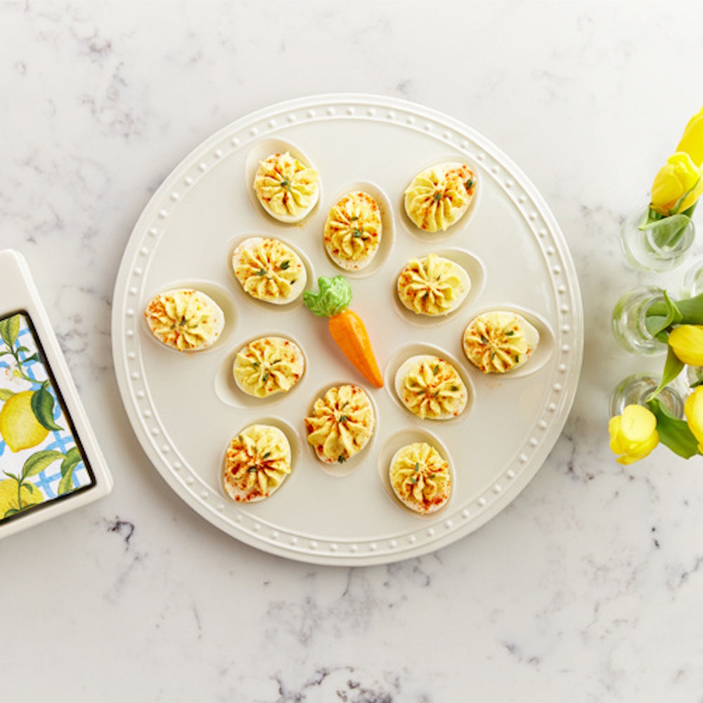 Deviled eggs on a white beaded ceramic platter with a ceramic carrot centerpiece and yellow tulips on a marble counter.