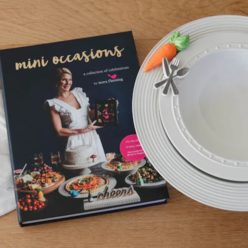 Mini Occasions cookbook by Nora Fleming next to a white ceramic plate with a carrot and utensil mini garnish.