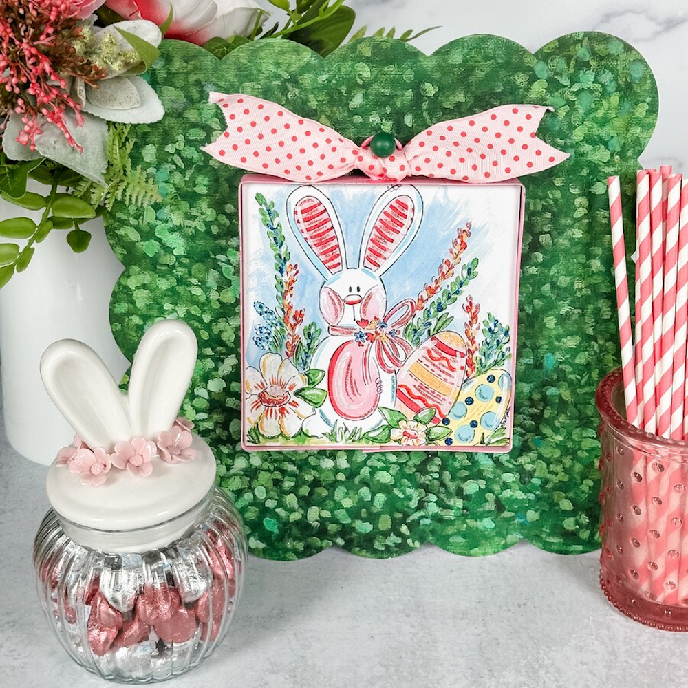 Easter bunny art with painted eggs and flowers on a green scalloped background with a pink polka dot bow.