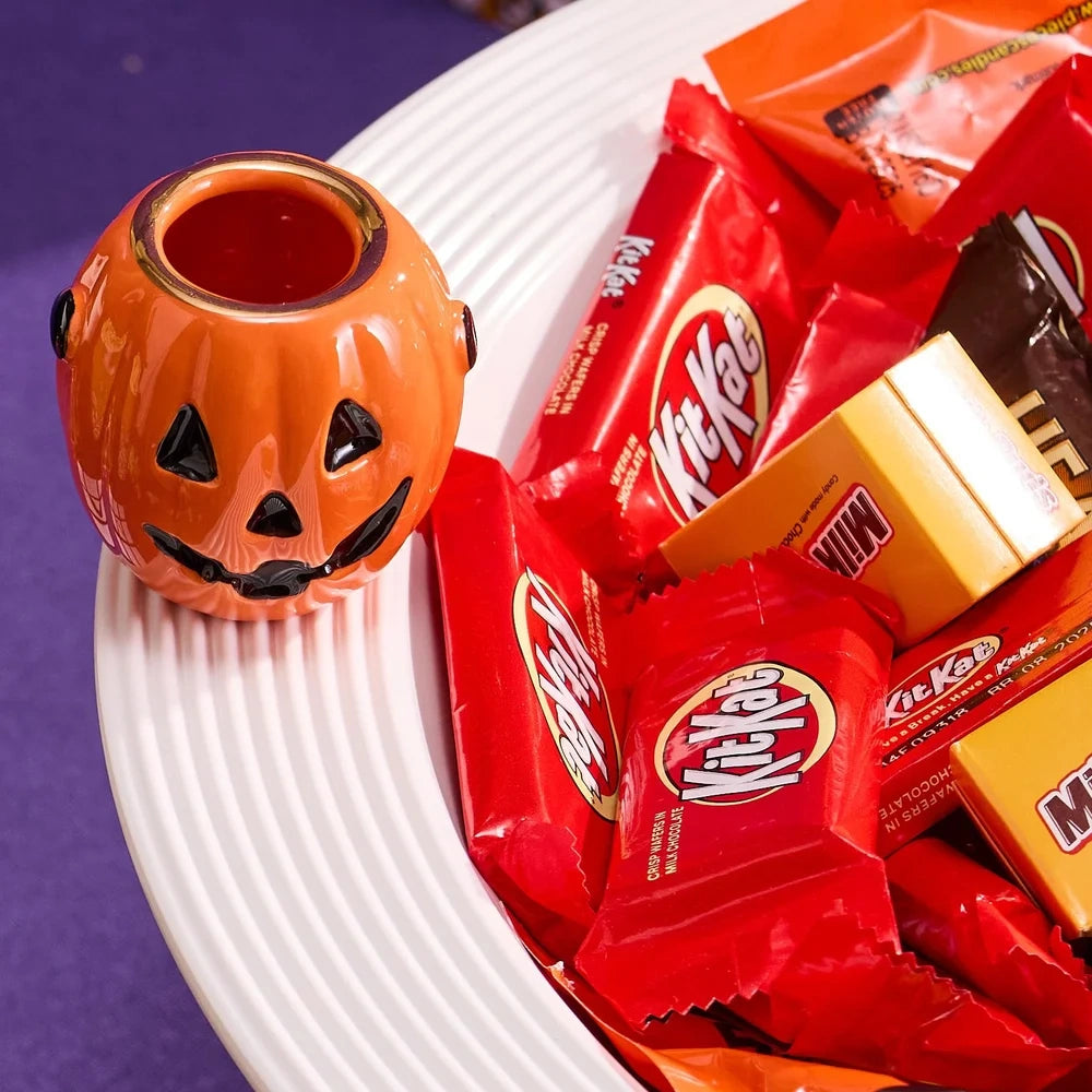 A small ceramic jack-o'-lantern holder sits on the rim of a white bowl filled with Halloween candy.