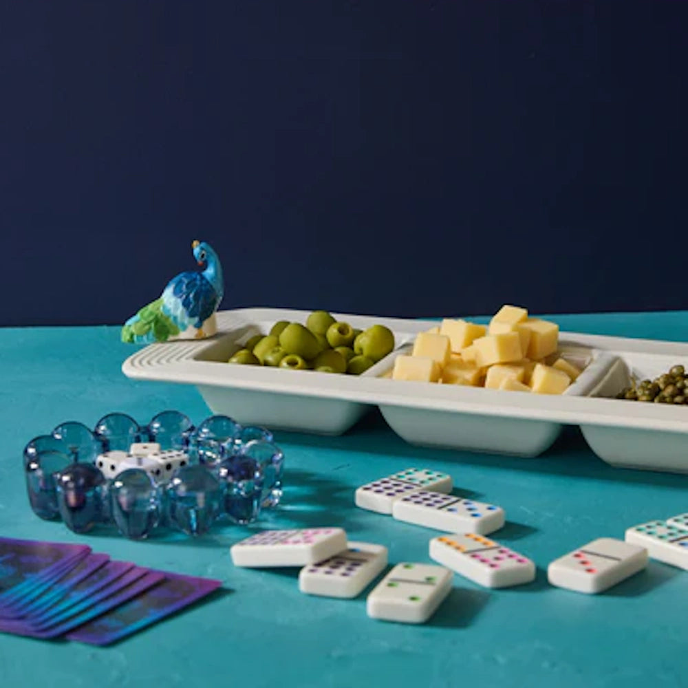 Peacock-themed appetizer tray with olives and cheese, alongside dominoes and cards on a blue surface.