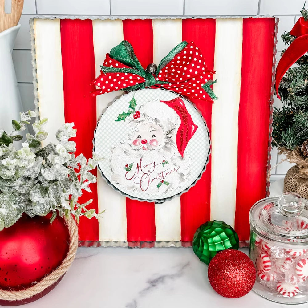 Front view of the round red and green Traditional Santa metal art plaque styled on a striped board with Christmas decor.