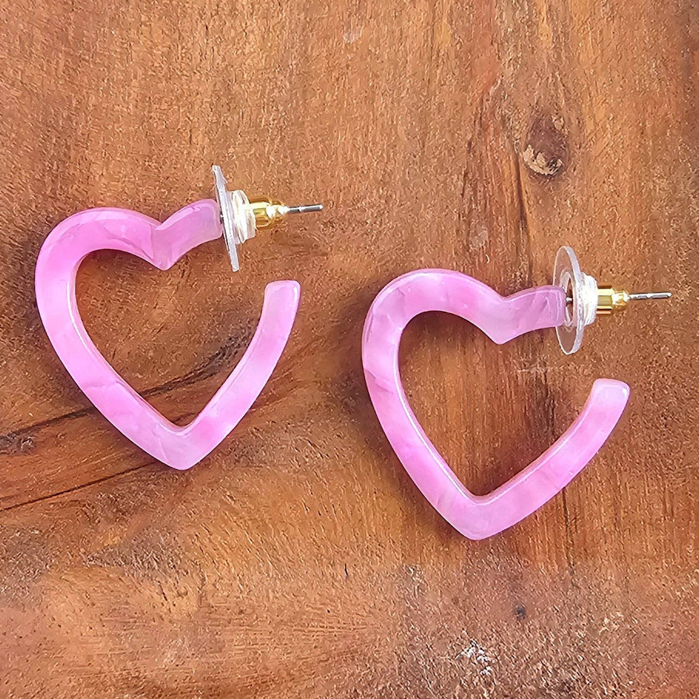Pink marbled resin mini heart-shaped hoop earrings with gold-toned posts held in a person's hand over a wooden surface.