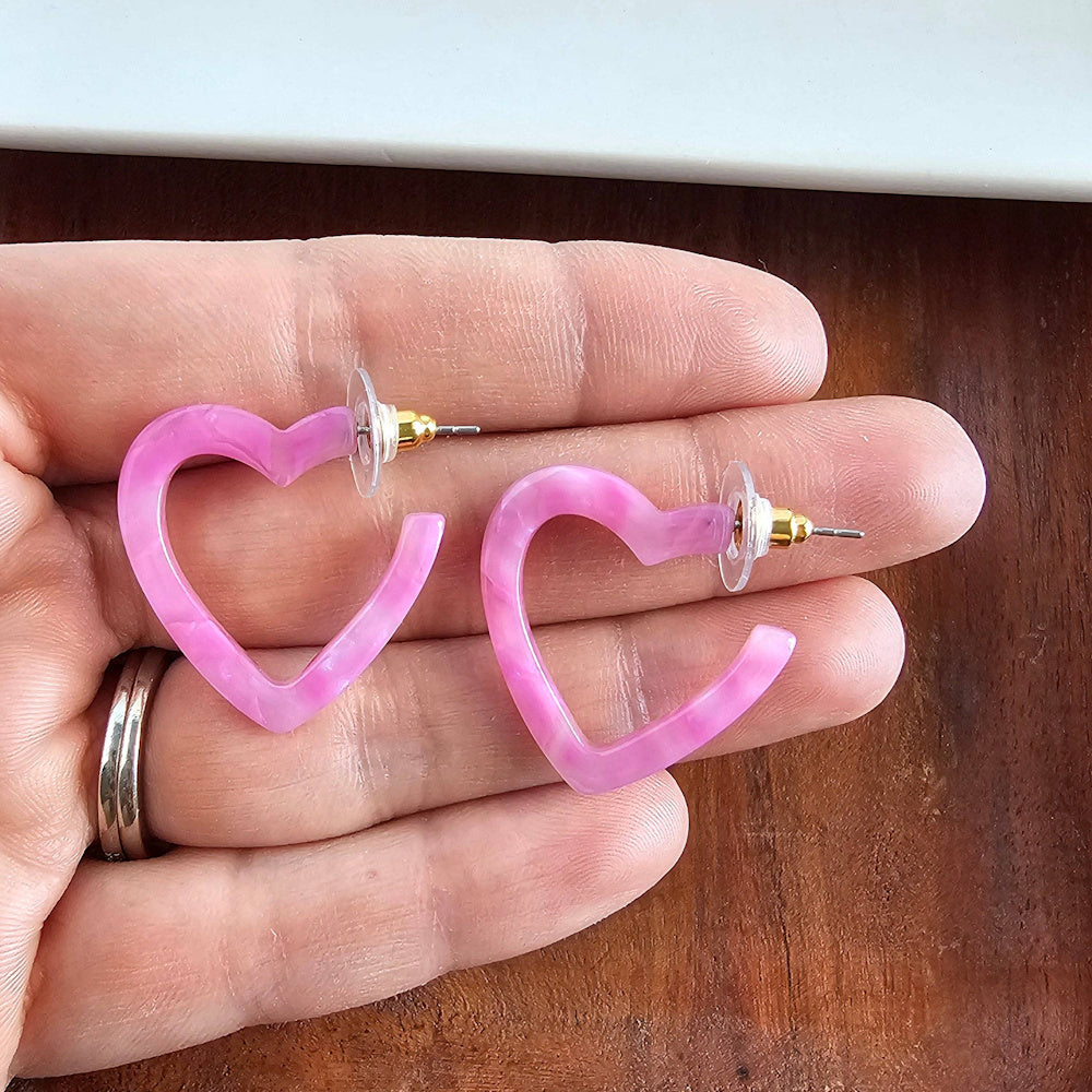 Pink marbled heart-shaped hoop earrings with gold-tone posts on a wooden background, perfect for a bubblegum bliss aesthetic.