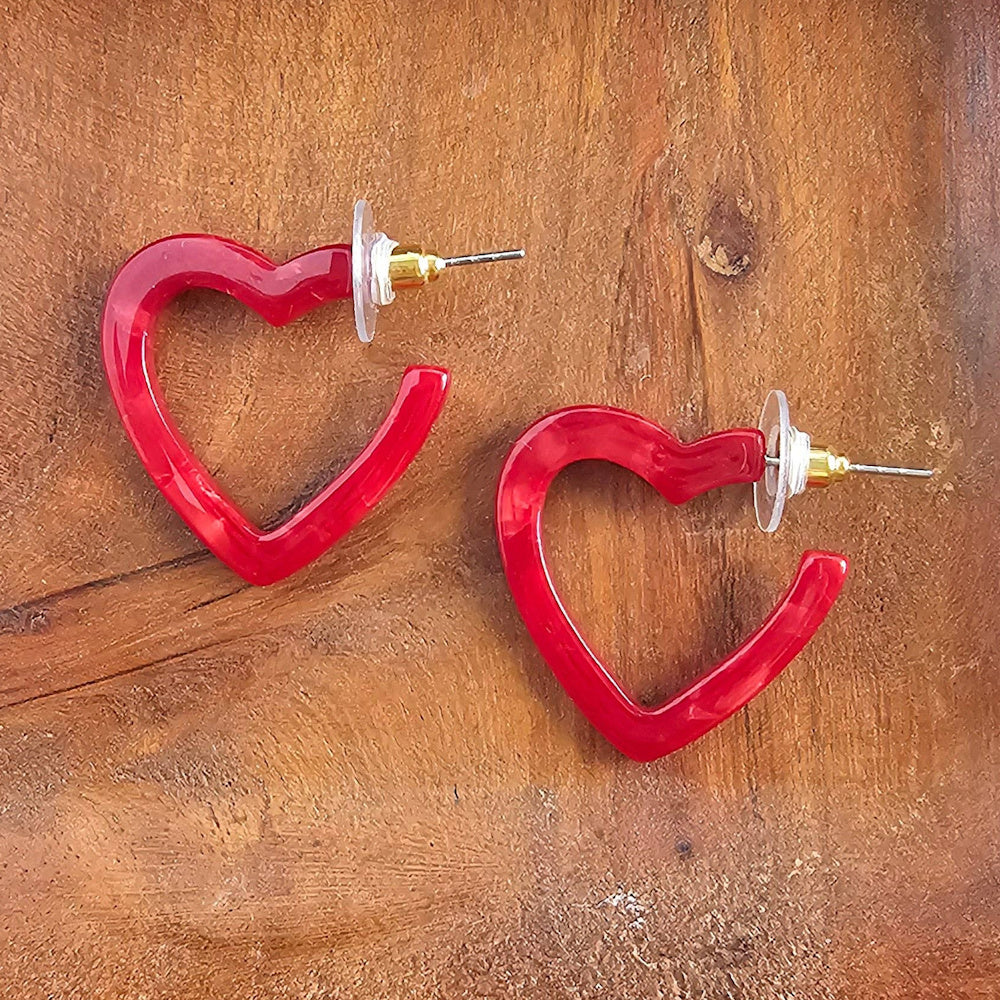 Pair of vibrant red acrylic heart-shaped hoop earrings with gold-toned posts on a textured wood background.