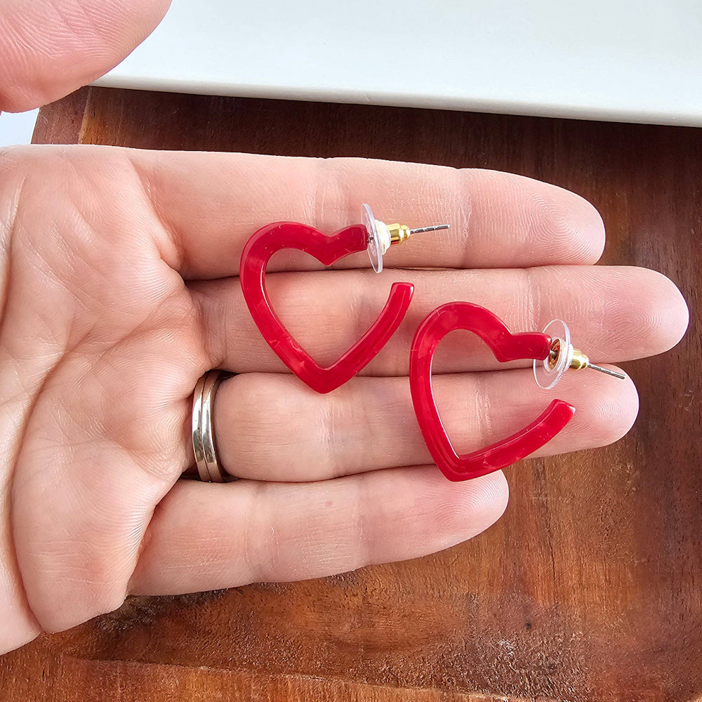 Pair of small red acrylic heart-shaped hoop earrings held in a hand over a wooden background.