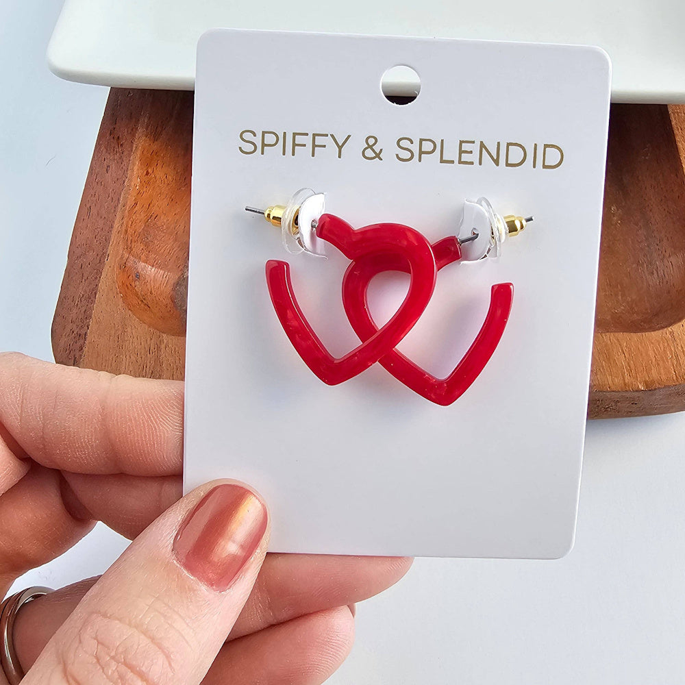Spiffy & Splendid red resin mini heart hoop earrings on a white display card held by a hand with rose gold nails.