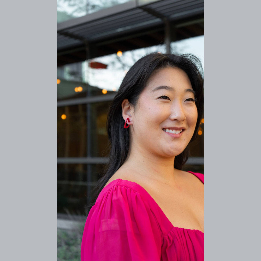 Smiling Asian woman wears a bright pink, puffed sleeve blouse and small red heart-shaped hoop earrings.