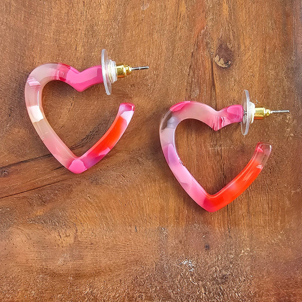 Pink marbled heart-shaped hoop earrings with gold-plated posts on a rustic wooden surface.