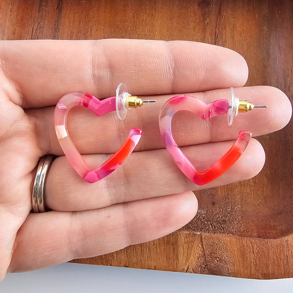 Mini heart-shaped acetate hoop earrings in marbled pink and red being held over a wooden background.
