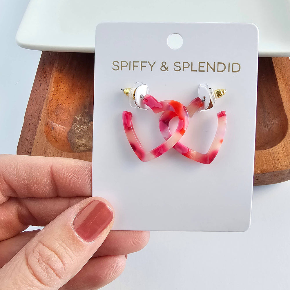 Spiffy & Splendid pink marbled acetate mini heart hoop earrings on a white display card held by a hand.