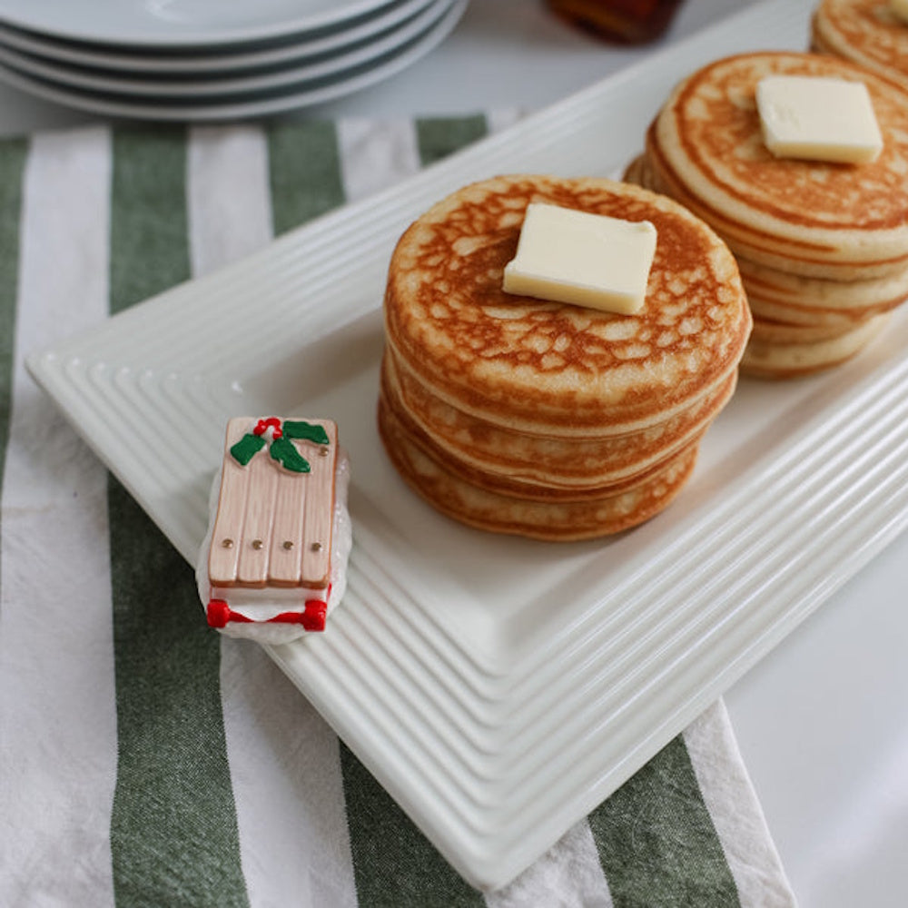 Stacks of golden pancakes topped with butter on a white platter beside a decorative mini Christmas sled.