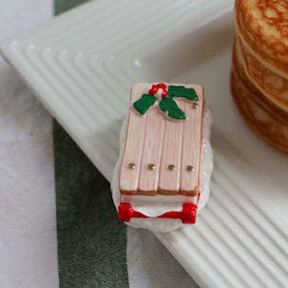 Small sled-shaped Christmas ornament with holly detail on a white textured plate next to a stack of pancakes.