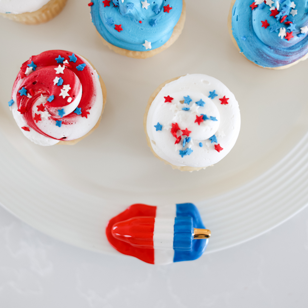 Red, white, and blue cupcakes with star sprinkles on a plate next to a melting rocket pop decoration.