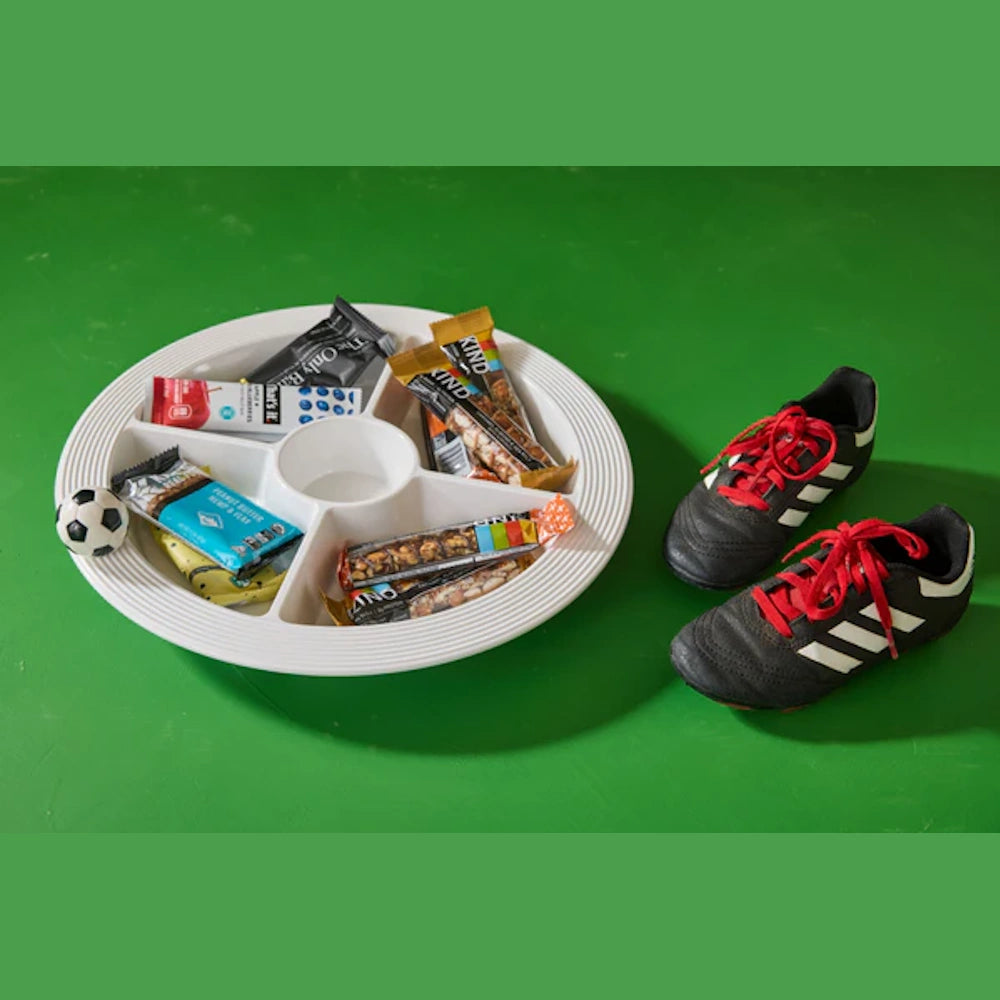 Soccer cleats next to a divided tray of snack bars and a mini soccer ball on a green background.