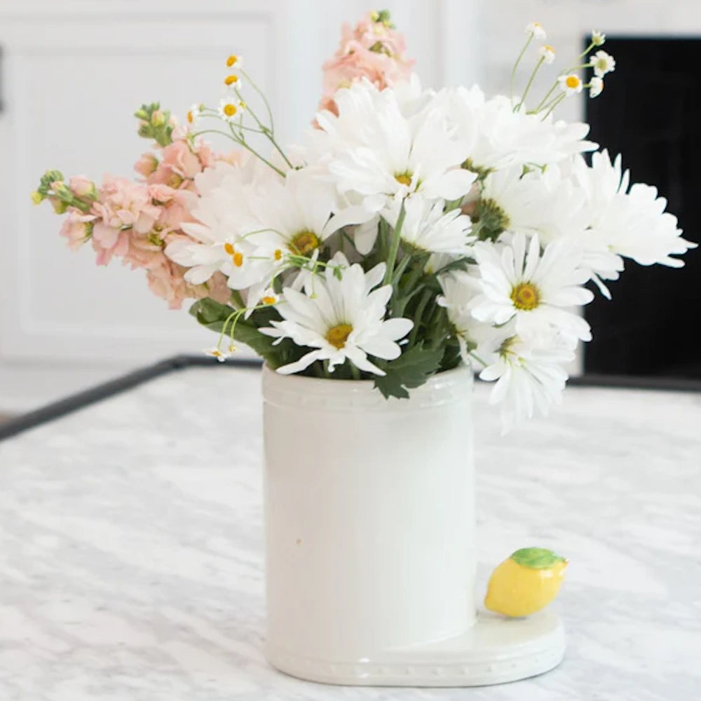 A bouquet of white daisies and pink flowers in a white ceramic vase with a small lemon detail on a countertop.