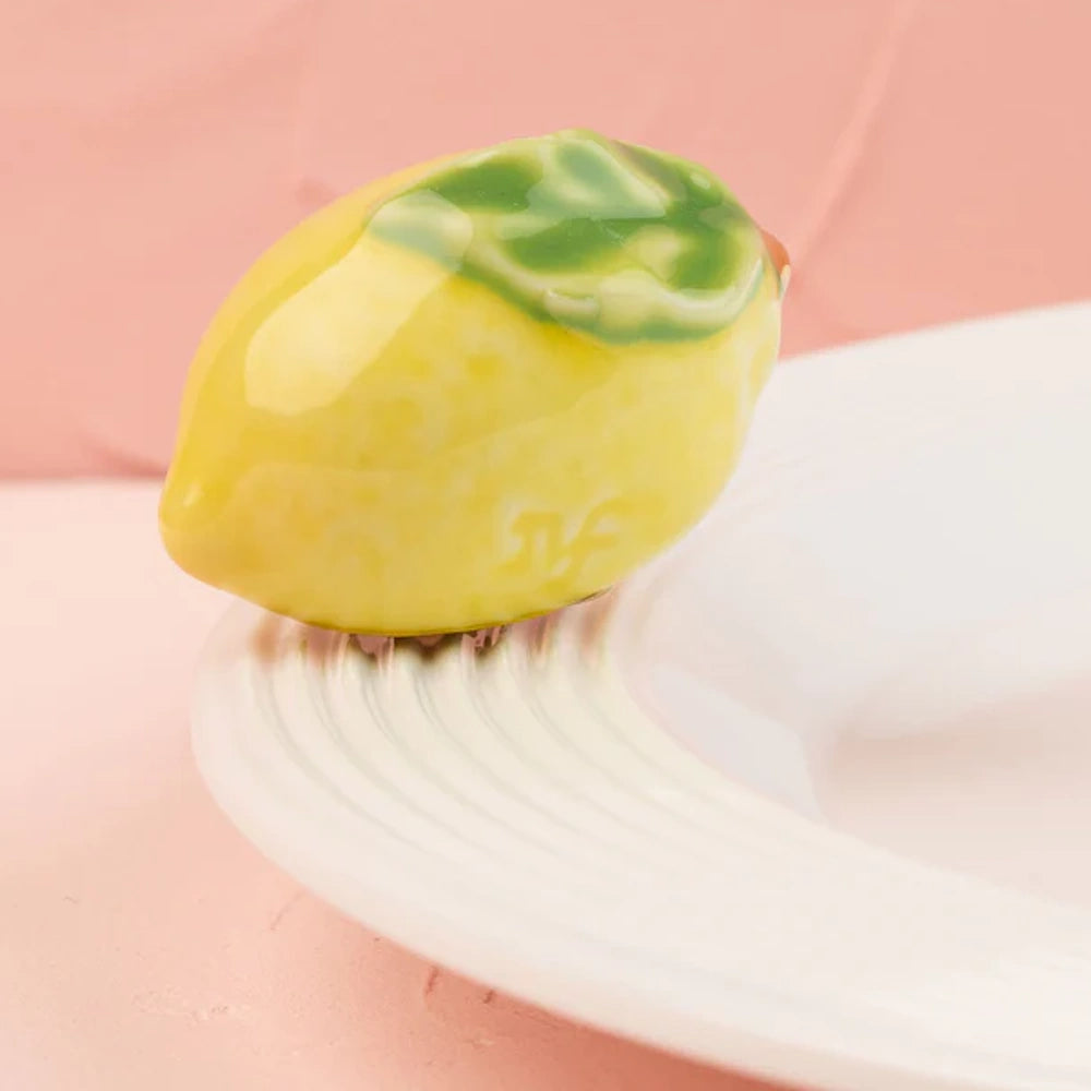 A small ceramic yellow lemon with a green leaf sitting on the rim of a white plate.