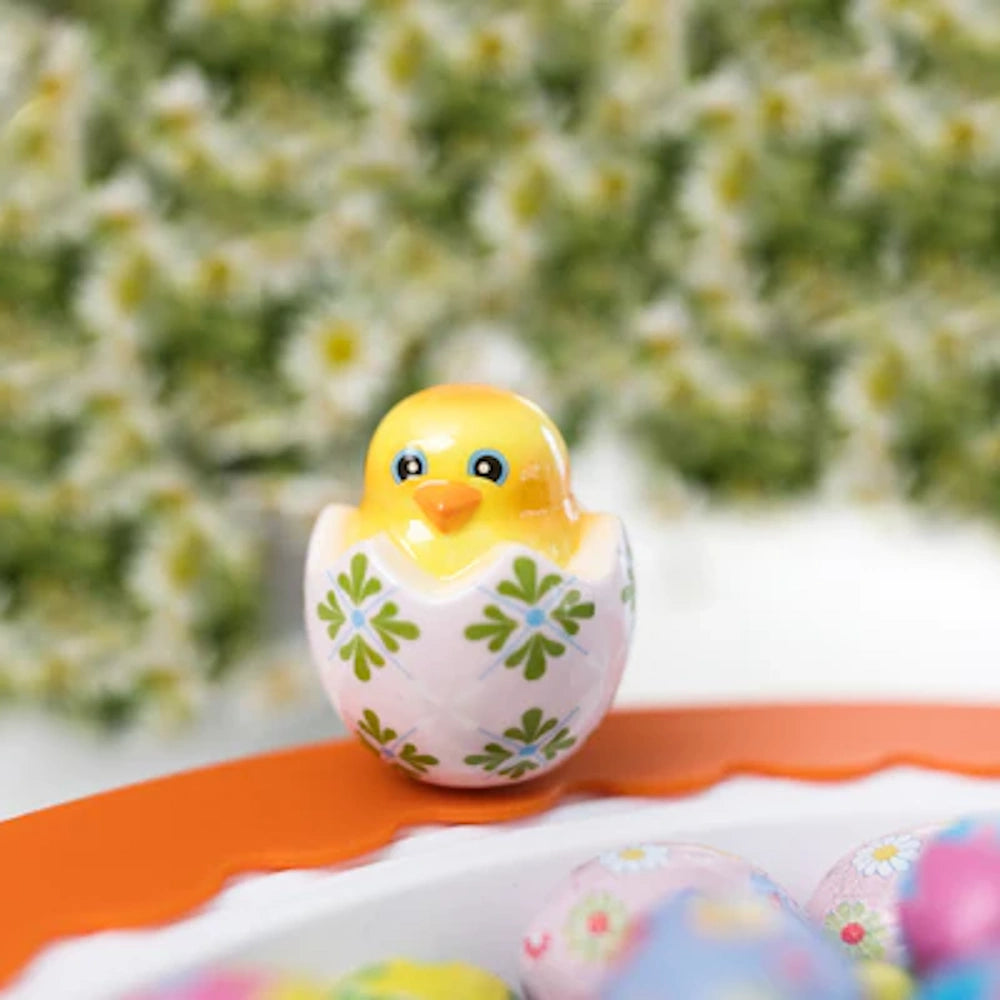 Yellow chick figurine hatching from a patterned Easter egg, set against a blurred floral background.