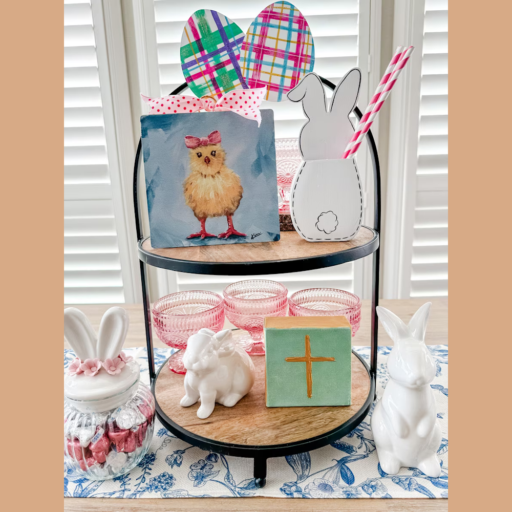 Two-tiered Easter display with a chick painting, bunny figurines, pink glass bowls, and a decorative cross on a wooden stand.
