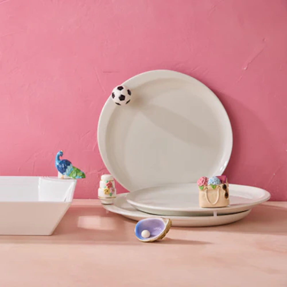 White ceramic plates and bowls decorated with small charms including a soccer ball and peacock against a pink background.