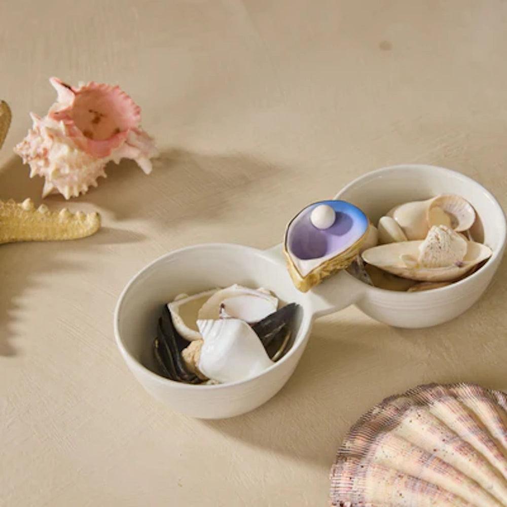 A decorative white double bowl filled with seashells and a pearl oyster figurine sits on a sandy surface.