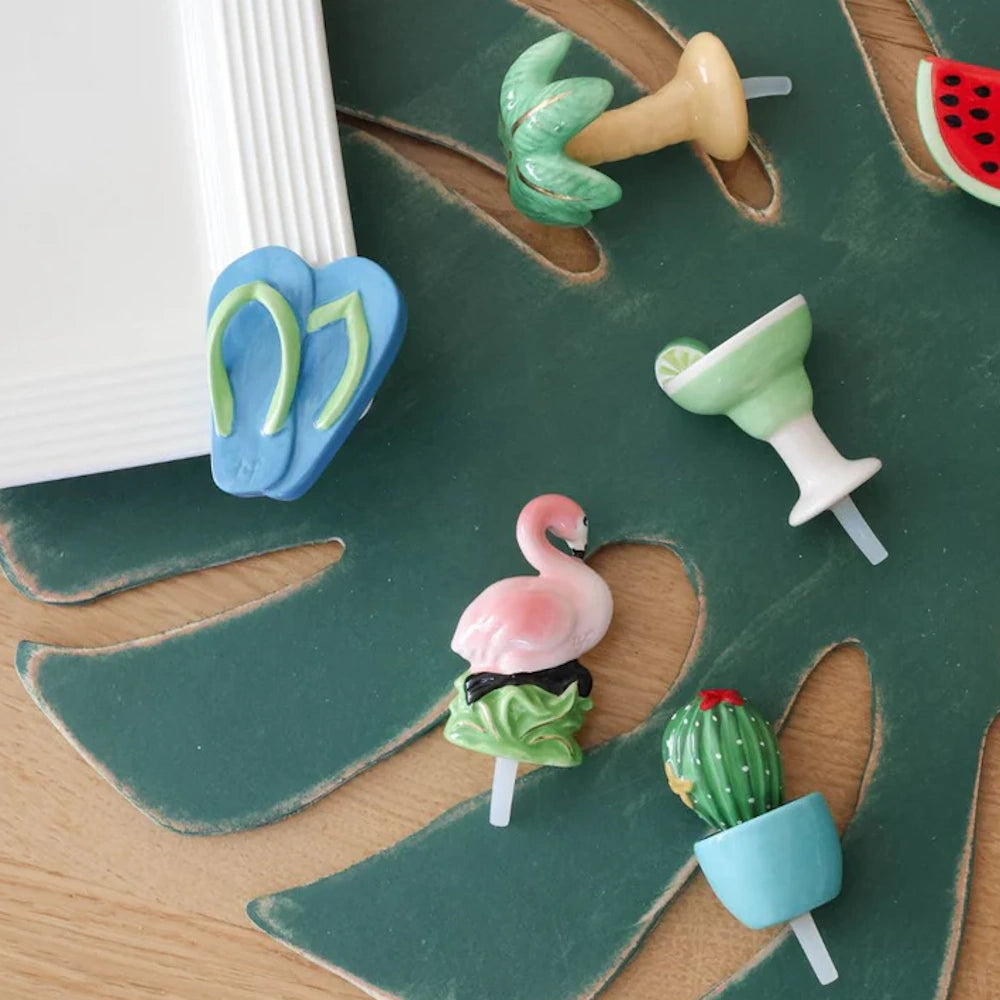 Tropical-themed ceramic drink markers including a flamingo, cactus, and palm tree on a green monstera leaf.