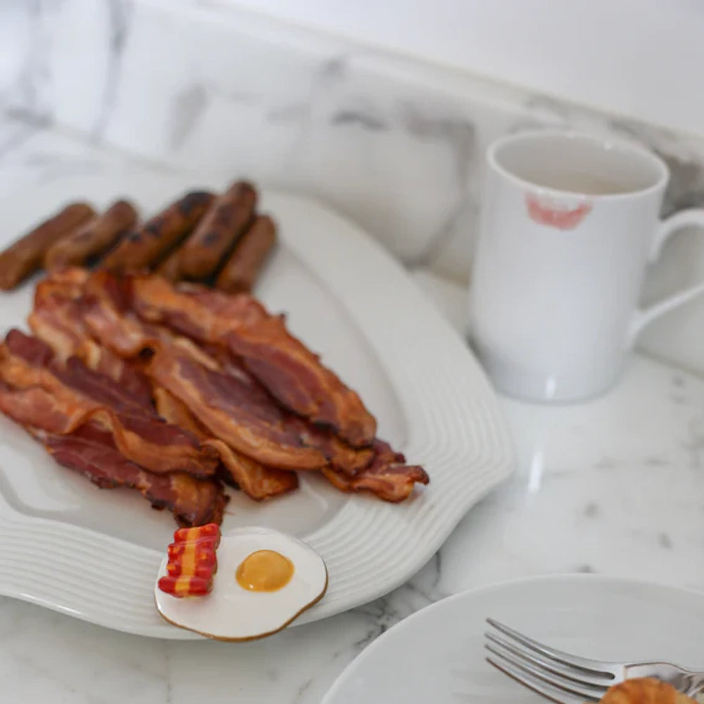 A plate of bacon and sausage next to a tiny miniature egg and bacon, beside a coffee mug with a lipstick mark.