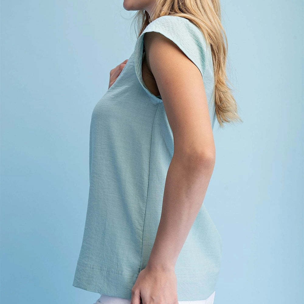 Side view of a woman wearing a mint V-neck blouse with trapunto stitching and loose cap sleeves against a blue backdrop.