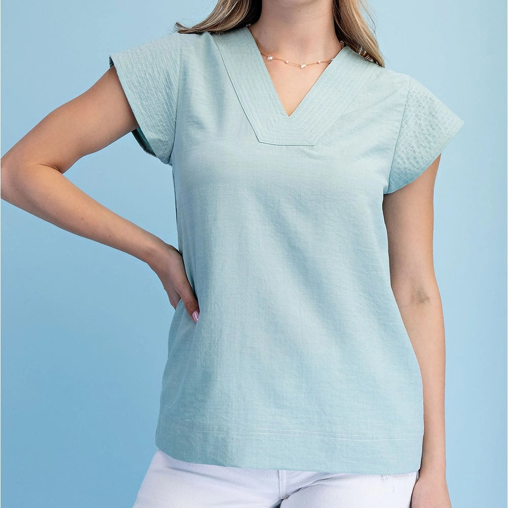 Mint trapunto stitched V-neck blouse with loose cap sleeves and white pants against a light blue background.