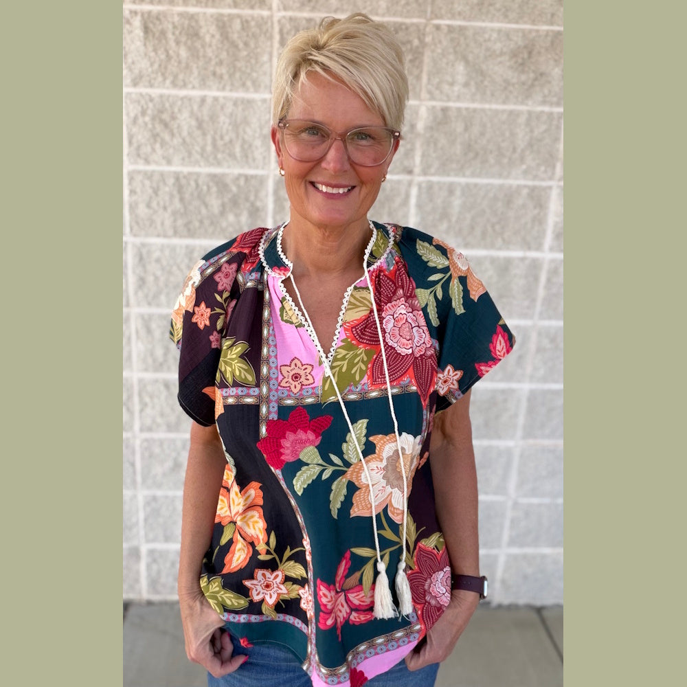 A woman with short blonde hair and glasses poses in a colorful, dark floral patchwork top with white tassel ties.