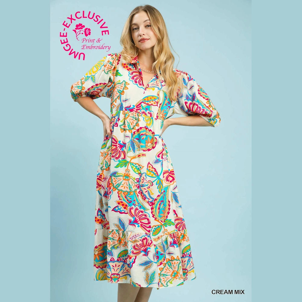 Woman wearing a cream maxi dress with colorful paisley print, puff sleeves, and a tiered hem against a blue background.