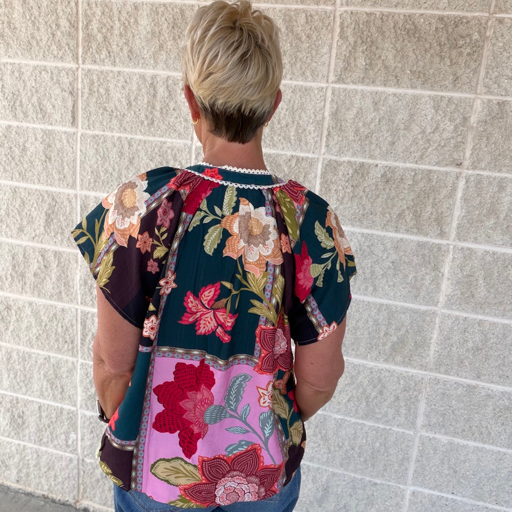 Back view of a woman wearing a mixed floral patchwork blouse with a lace-trimmed neckline against a stone wall.