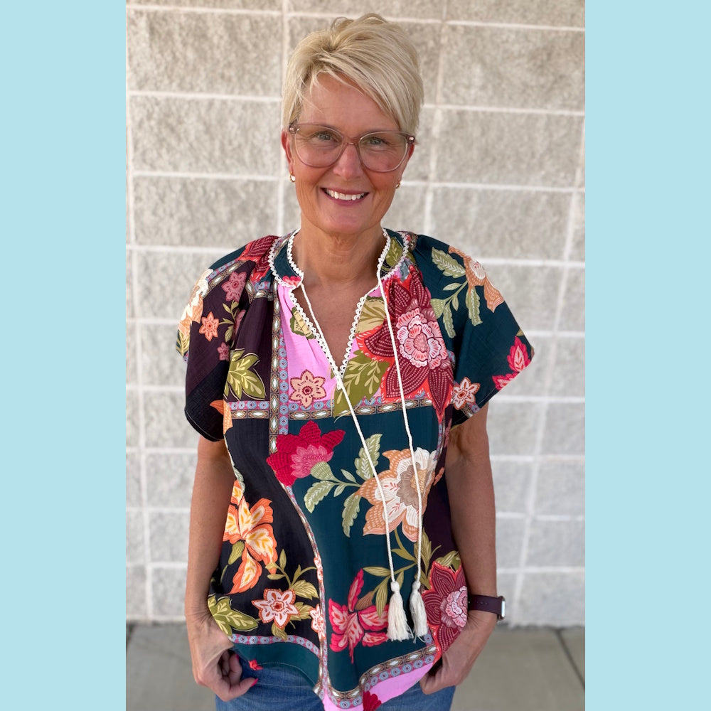 Woman wearing a floral patchwork tassel-tie top with a v-neck and white lace trim against a white brick wall.