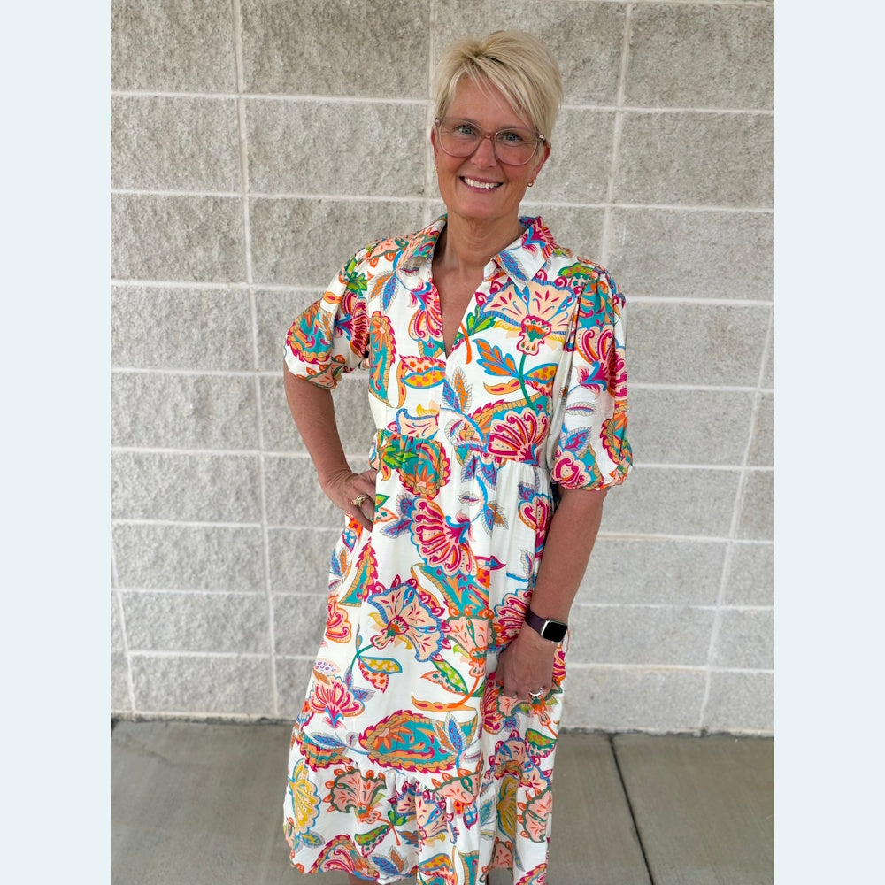 Woman in a colorful paisley print maxi dress with puff sleeves and a tiered skirt standing against a white brick wall.
