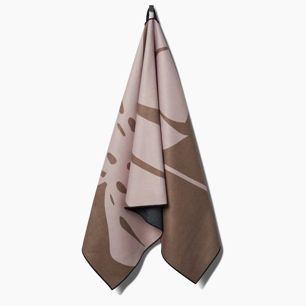 A draped beach towel hanging from a hook, featuring a large monstera leaf pattern in pink and brown.